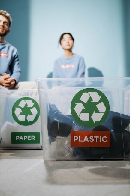 Close-up view of two transparent recycling bins placed on a table, one labeled 'PAPER' with a green recycling symbol and the other labeled 'PLASTIC' with a green recycling symbol, indicating waste sep