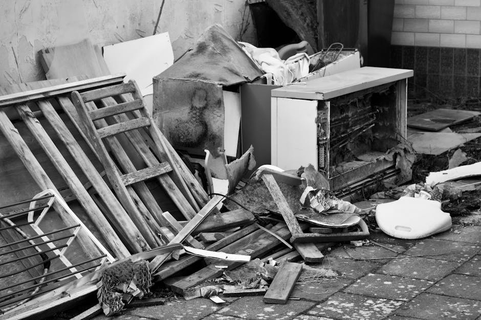 A pile of discarded household items and furniture outside a property, including a wooden step ladder leaning against a wall, various broken wood pieces, a damaged cabinet with its door ajar, a plastic container, and pieces of cardboard and packaging materials scattered on a paved ground. In the background, part of a brick and concrete wall is visible, with some fabric and wrapping materials draped over the furniture. The scene depicts the aftermath of furniture disposal as part of a home relocation or clearing process, with some items possibly ready for collection by removal services. The overall setting is outdoors, with natural lighting illuminating the cluttered scene, aligning with professional removals and moving logistics described by Movers Kingston upon Thames.