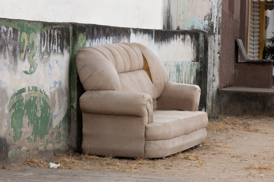 A beige upholstered armchair with visible wear and tear, including a torn section on the backrest displaying yellowed foam, is positioned outside against a weathered concrete wall with peeling paint and faded graffiti in green and blue. The area around the chair has dirt and scattered leaves, indicating it is placed on a rough, uneven pavement or ground. To the right, a dark brown concrete staircase leads up to a door, and a section of a wooden window shutter is partially visible. The scene appears to be part of a house relocation or furniture disposal process, with the chair temporarily placed outdoors, possibly awaiting removal by a professional moving service like Movers Kingston upon Thames.