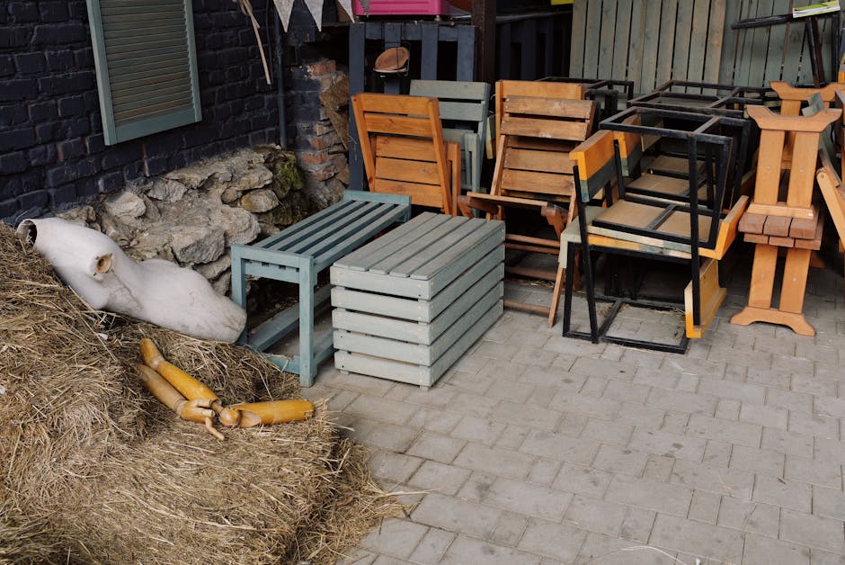 A pile of discarded household items and furniture outside a property, including a wooden step ladder leaning against a wall, various broken wood pieces, a damaged cabinet with its door ajar, a plastic container, and pieces of cardboard and packaging materials scattered on a paved ground. In the background, part of a brick and concrete wall is visible, with some fabric and wrapping materials draped over the furniture. The scene depicts the aftermath of furniture disposal as part of a home relocation or clearing process, with some items possibly ready for collection by removal services. The overall setting is outdoors, with natural lighting illuminating the cluttered scene, aligning with professional removals and moving logistics described by Movers Kingston upon Thames.