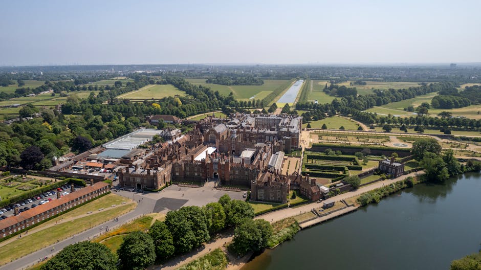 An aerial view of Hampton Court Palace situated near the River Thames, showcasing historic brick and stone architecture with ornate turrets, large windows, and detailed facades. The palace is surrounded by well-maintained gardens, manicured lawns, and symmetrical hedge layouts, with pathways visible among the greenery. Beyond the palace, expansive fields and wooded areas stretch into the distance, with a long straight driveway leading from the palace entrance into the surrounding countryside. The river runs along the edge of the property, with a walkway and some boats moored nearby. The sky is clear, indicating good weather, and the overall scene emphasizes the grandeur and historic significance of the site, relevant to house removals, relocation services, and furniture transport for large antiques and valuable items as part of a moving process coordinated by Movers Kingston upon Thames for the PAGE_TITLE.