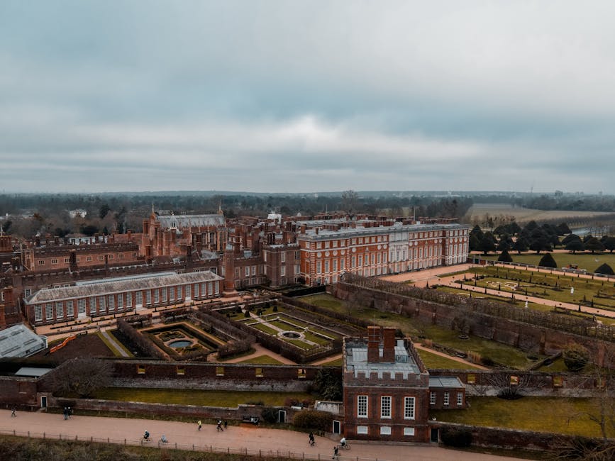 An aerial view of Hampton Court Palace showing its historic red brick architecture, with multiple connected buildings featuring ornate gables, chimneys, and large windows. Surrounding the palace are formal gardens with neatly trimmed hedges, gravel paths, and grassy lawns, some with geometric patterns and water features. The gardens are enclosed by low brick walls, and a pathway along the perimeter is visible with people walking. Overcast skies cast diffuse lighting over the scene. This setting is part of a house removals project by Movers Kingston upon Thames, involved in packing and moving large antiques and furniture from the palace, with some objects likely being loaded onto a van positioned nearby, outside the property, for transportation as part of the logistics for the KINGSTON UPON THAMES move.