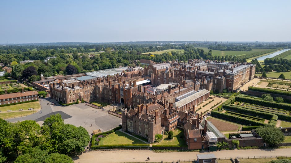 An aerial view of Hampton Court Palace situated near the River Thames, showcasing historic brick and stone architecture with ornate turrets, large windows, and detailed facades. The palace is surrounded by well-maintained gardens, manicured lawns, and symmetrical hedge layouts, with pathways visible among the greenery. Beyond the palace, expansive fields and wooded areas stretch into the distance, with a long straight driveway leading from the palace entrance into the surrounding countryside. The river runs along the edge of the property, with a walkway and some boats moored nearby. The sky is clear, indicating good weather, and the overall scene emphasizes the grandeur and historic significance of the site, relevant to house removals, relocation services, and furniture transport for large antiques and valuable items as part of a moving process coordinated by Movers Kingston upon Thames for the PAGE_TITLE.