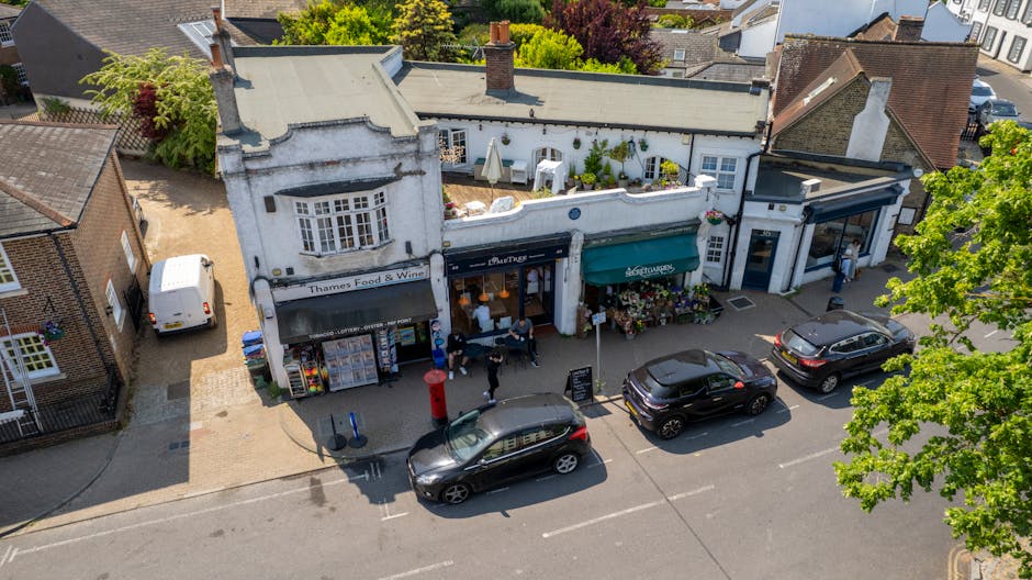 An aerial view of a street in Kingston upon Thames showing a small commercial building with a white facade and black signage, housing a food and wine shop. In front of the shop, there are several parked cars including a black hatchback, a black SUV, and another dark vehicle, with a red postal box near the curb. The sidewalk features a few pedestrians, some entering or leaving the shop, and a small display of potted plants and flowers outside. The scene captures the busy environment during daytime, with natural sunlight illuminating the storefronts, vehicles, and neighboring buildings, reflecting typical urban activity related to moving or deliveries in a commercial area. This visual aligns with the context of house or business relocations, highlighting the logistics and shopfront access involved, similar to services offered by Movers Kingston upon Thames for commercial and household removals.