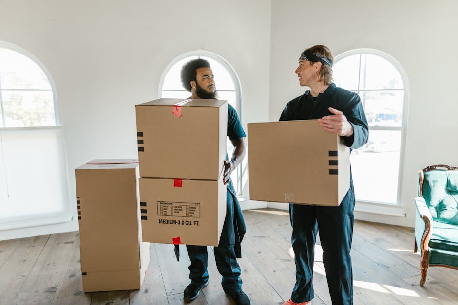 Two removal movers from Movers Kingston upon Thames are inside a bright, well-lit room with white walls and three large arched windows. They are engaged in a home relocation process, each holding a medium-sized cardboard box with printed labels, black tape, and red markers. The mover on the left, who has a beard and curly hair, is carrying two stacked boxes, while the other, wearing a headband and casual clothing, is preparing to load or hand over a box. Several other cardboard boxes, some sealed with tape and labels, are placed on the light wooden floor, indicating packing and moving preparations. The room's environment includes a vintage green armchair with wooden accents on the right side of the image, and the natural daylight streaming through the windows enhances visibility, highlighting the furniture, packing materials, and the active loading process involved in a professional furniture transport or house removal operation.