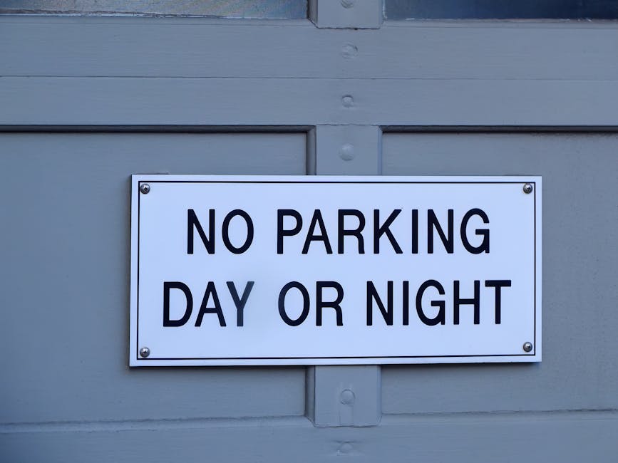 A rectangular white sign with black bold lettering reading 'NO PARKING DAY OR NIGHT' is affixed to a grey painted wooden door or paneling. The sign is mounted with four visible screws, one at each corner. The background consists of grey wood with horizontal and vertical panel details, indicating an exterior door or garage. The photograph appears to be taken during daylight, with natural lighting illuminating the sign and wooden surface. This signage may relate to parking restrictions relevant to house removals or moving services offered by Movers Kingston upon Thames, who coordinate logistics for home relocations within the Kingston upon Thames area, including compliance with local parking permit rules for loading and unloading a vehicle outside a property.