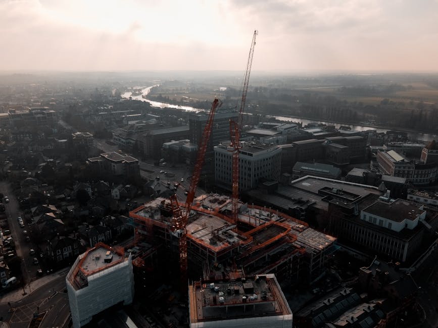 Aerial view of a cityscape during daytime showing a large construction site with two tall orange cranes actively positioned over a multi-story building under construction. The building's concrete framework and partially completed floors are visible, covered with temporary safety barriers and construction materials such as wooden planks and metal supports. Surrounding the site, a mix of completed office buildings, residential structures, and streets with parked cars are seen, extending towards a river in the background. The scene is illuminated by natural daylight, with a hazy sky overhead, illustrating an ongoing home relocation or building development process supported by construction equipment and urban infrastructure. This image is relevant to house removals, moving logistics, and construction activities in Kingston upon Thames, with [COMPANY_NAME] involved in logistics or building site services related to packing and moving.