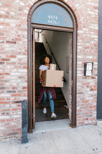 A woman with curly hair, dressed in casual clothing including a white t-shirt, blue jeans, and white sneakers, is seen walking through the entrance of a brick building while carrying two cardboard boxes, one of which appears to be sealed with packing tape. Behind her, internal stairs with a dark handrail are visible, leading upwards inside the property. Someone else can be seen at the top of the stairs, holding another box and assisting with the home relocation process. The building has a gently arched doorway with a glass window above the door displaying the numbers '4474' and '4476', and an intercom system fixed beside the entrance on the right. The paving outside is clean concrete, and the scene is well-lit, indicating daytime. This image captures the process of packing and moving items during a residential relocation, with a focus on the coordinated effort involved in short-distance furniture transport within an urban property, aligning with house removals services provided by Movers Kingston upon Thames.