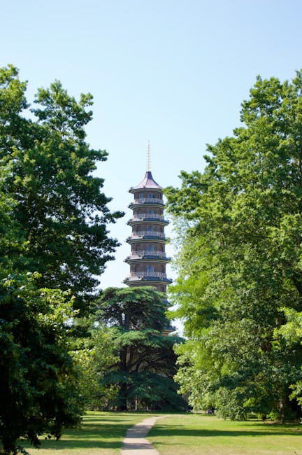 A tall, multi-tiered pagoda-style tower with a pointed spire at the top, situated in a green park environment. The tower is surrounded by lush deciduous trees with vibrant green foliage, some partially obscuring the lower levels of the structure. The sky is clear and light blue, indicating a bright, sunny day. In the foreground, a paved pathway runs through the grassy area, leading towards the tower, which appears to be a historic or cultural landmark. The scene is tranquil and well-maintained, with the tower serving as a focal point amidst the landscaped surroundings, illustrating a typical setting for house removals or relocation services operating in the Kingston upon Thames area, as highlighted on the website moverskingstonuponthames.co.uk.