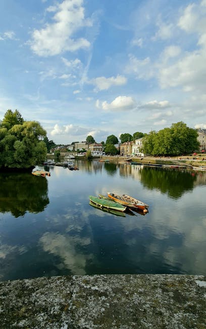 A tranquil river scene captured during daylight, featuring calm waters reflecting partly cloudy blue sky and scattered white clouds above. Along the riverbank, there are several boats secured to the shoreline, including a wooden rowing boat and a small motorboat, positioned near a concrete or stone edge. In the background, there are residential buildings with light-colored facades and a variety of mature green trees lining the opposite bank, providing a peaceful setting. The image perspective appears to be from a vantage point close to the water's edge, possibly on a viewing platform or a walkway, with natural lighting highlighting the serene ambiance. This scene illustrates a quiet home relocation or furniture transport setting connected to moving services, where the environment complements logistical activities such as loading or unloading nearby, supported by the presence of boats and natural surroundings, aligning with the context of same-day removals and local house moving in Kingston upon Thames.
