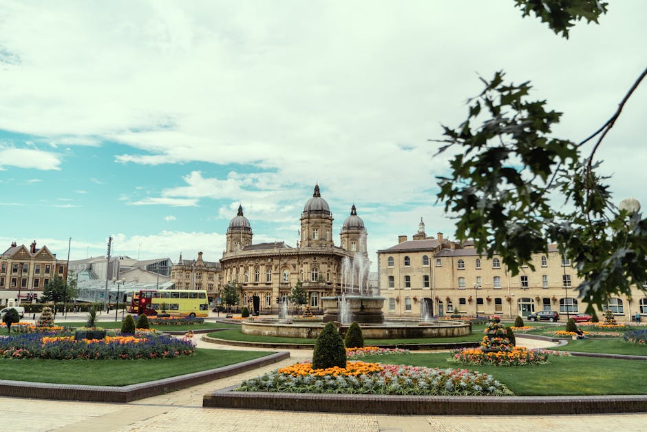 A wide exterior view of a city square in Kingston upon Thames showcasing a large ornate building with domed towers and classical architectural features, situated behind a fountain with multiple streams of water. The foreground features well-maintained flowerbeds with colorful flowers, trimmed bushes, and paved pathways. A double-decker bus is visible on the street adjacent to the square, while a red van and parked cars are seen nearby. A tree branch with green leaves extends into the top right corner of the image, partially framing the scene. The sky above is partly cloudy with patches of blue visible, and the environment suggests a busy, scenic location suitable for area-based house removals or furniture transport, with the open space and urban setting indicating a typical town centre atmosphere. This image effectively captures the setting for local moving or relocation services, aligning with the page offering same-day removals in Kingston upon Thames from Movers Kingston upon Thames.
