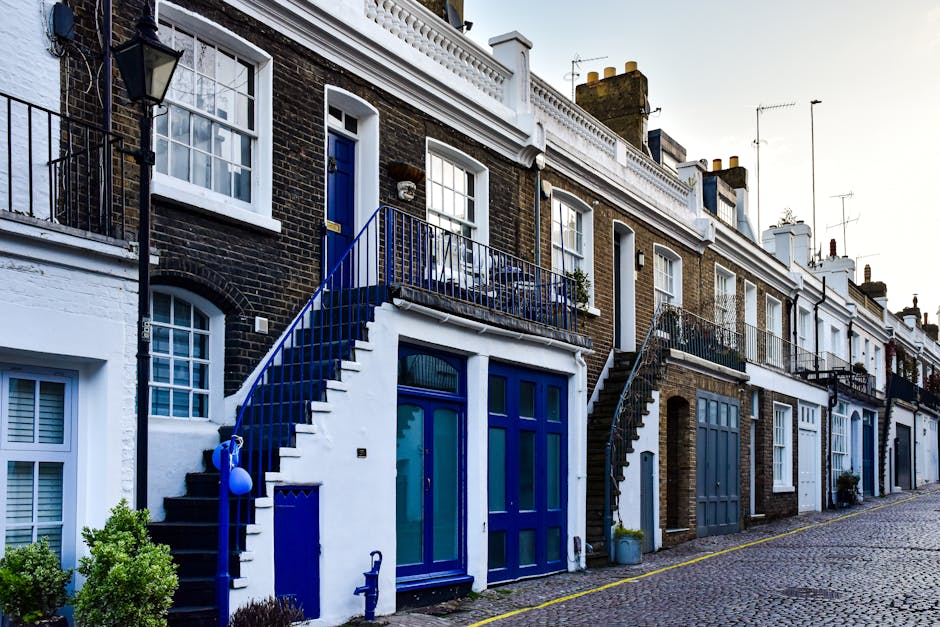 A white moving van with a closed cargo area is parked on a narrow street in Kingston upon Thames during daytime. The van is positioned close to a multi-storey residential building with contemporary wi